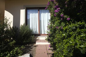 A pathway leading to a door with a white curtain, surrounded by greenery and shrubs.