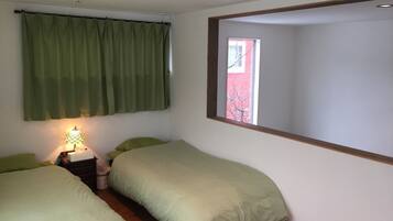 A bedroom with two single beds, a nightstand with a lamp, a window with a view of a red building, and a curtain.