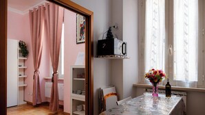 A room with a wooden floor, a dining table with a floral tablecloth, a vase with flowers, a radiator, and a window with sheer curtains. - Wonderful beautiful apartment in the heart of Rome (Esquilino Colosseum area) (Roma)