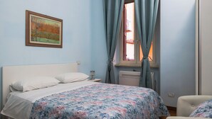A bedroom with a bed, a nightstand, a window with curtains, a radiator, and a painting on the wall. - Wonderful beautiful apartment in the heart of Rome (Esquilino Colosseum area) (Roma)