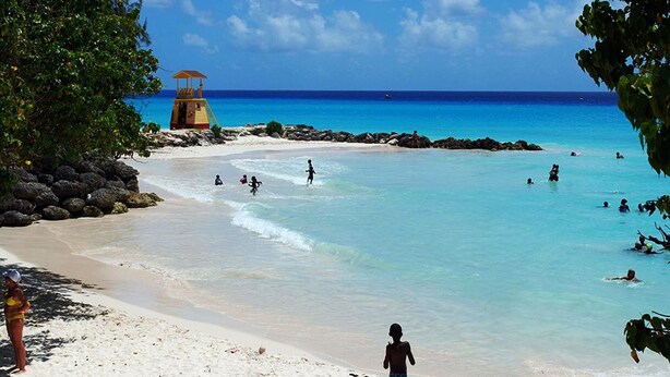 Een strand met helder turquoise water, een uitkijktoren van de badmeester en mensen die zwemmen en over het zand wandelen.