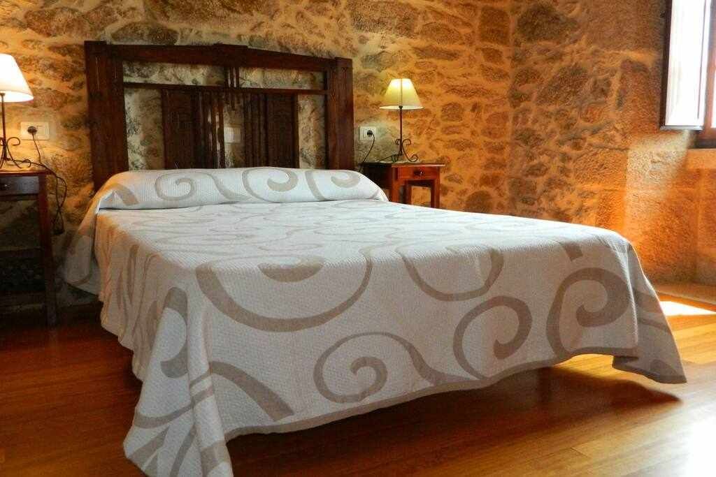 Een slaapkamer met een stenen muur, een houten hoofdbord, een bed met een geborduurde spreideken, twee nachtkastjes met lampen, en een raam aan de rechterkant.