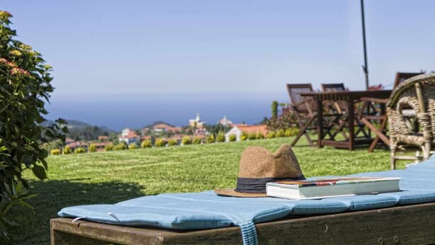 A wooden bench with a hat and book, a wicker chair, and an umbrella in a grassy outdoor area with a view of the sea and buildings in the background. - THE ERA OF SOMAO (Somado)