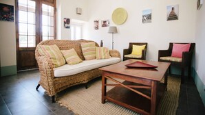 Una sala de estar con un sofá de mimbre, una mesa de centro de madera y dos sillones. Hay cuadros colgados en la pared y una lámpara.