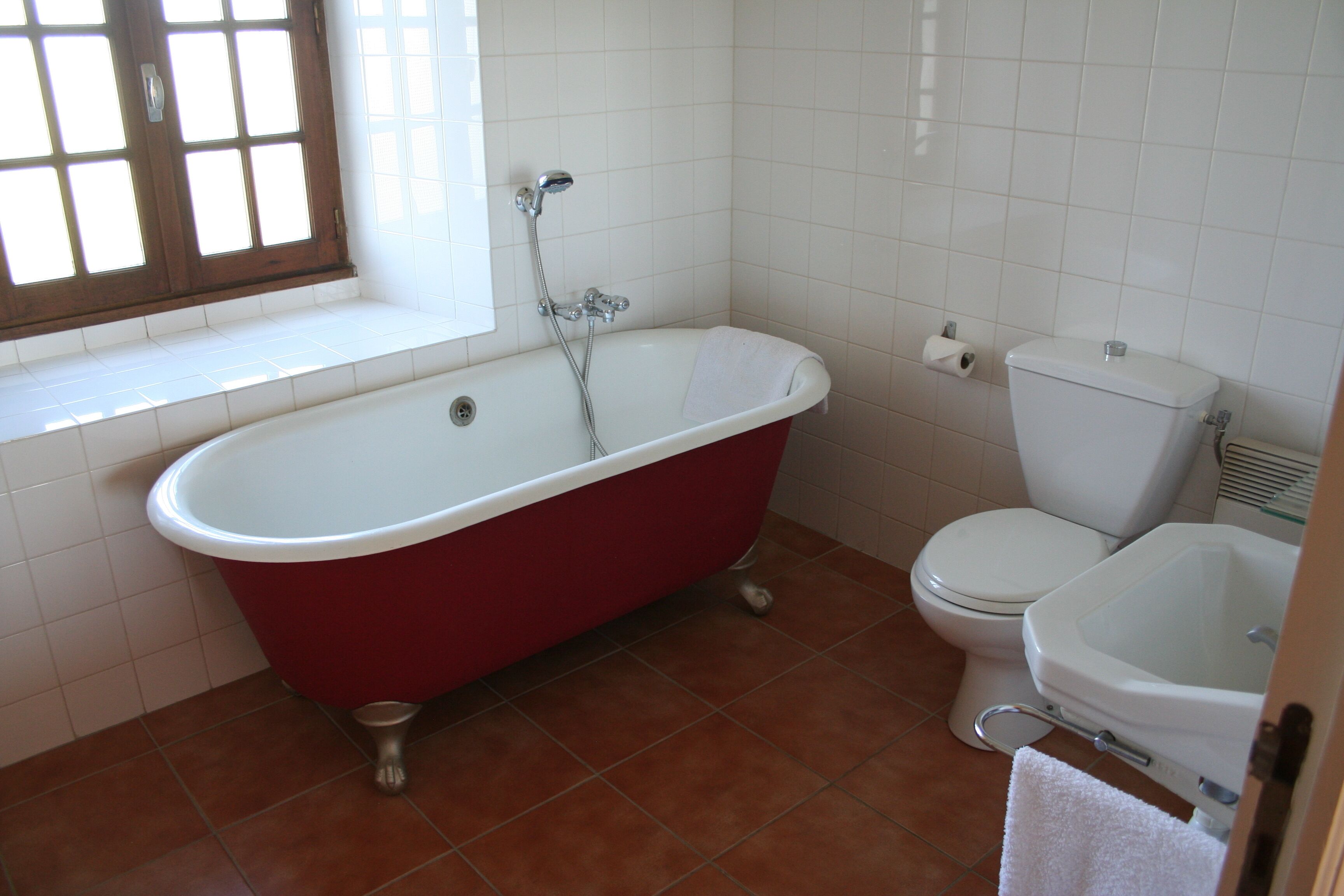 Un baño con una bañera roja tipo pata de león, un inodoro blanco y un lavabo pedestal blanco.
