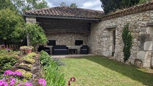 Un edificio de piedra con techo de tejas, una pared de piedra y un patio con césped, plantas en macetas y flores.