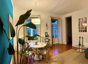 A dining area with a round table and chairs, a hanging light fixture, and large green plants. - Spirit of Time - Elegant 2-Bedroom Retreat with Garden Views (Lisboa)