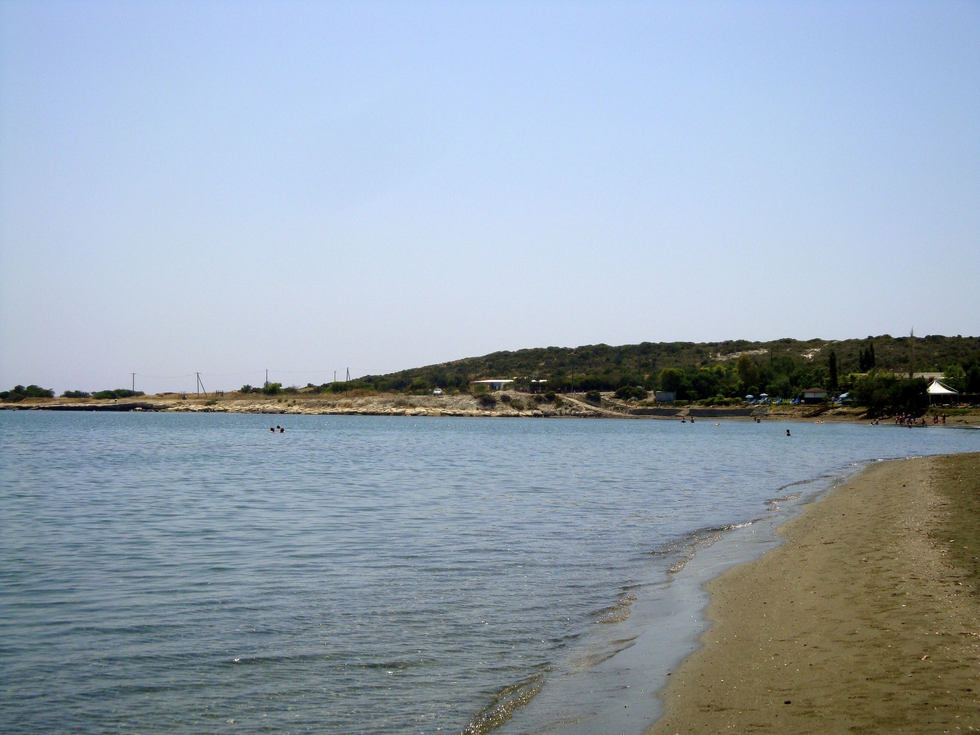 A beach with calm water, a sandy shore, and a hilly landscape in the background.