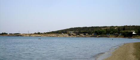 A beach with calm water, a sandy shore, and a hilly landscape in the background.