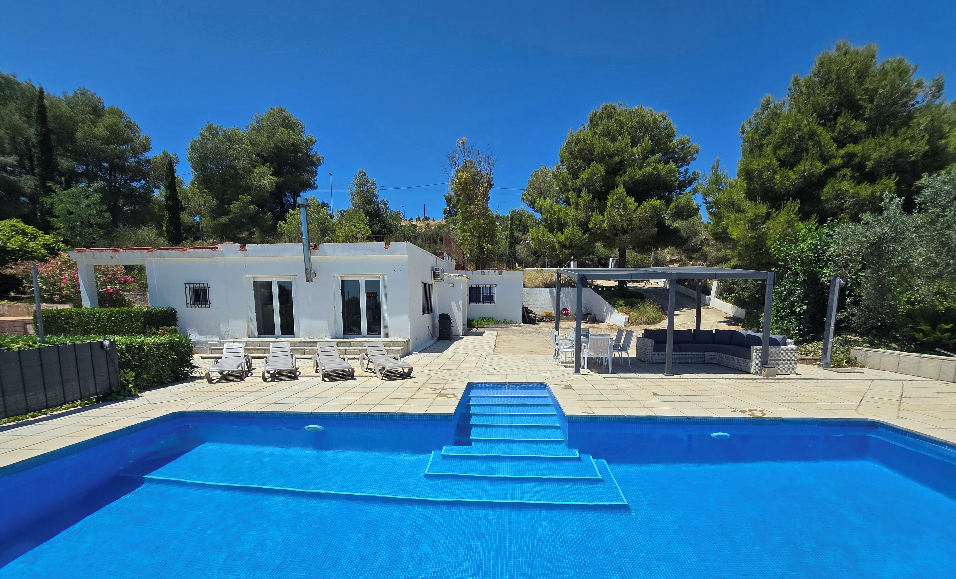 A swimming pool with steps leading into it, surrounded by a patio area with lounge chairs and a covered seating area.