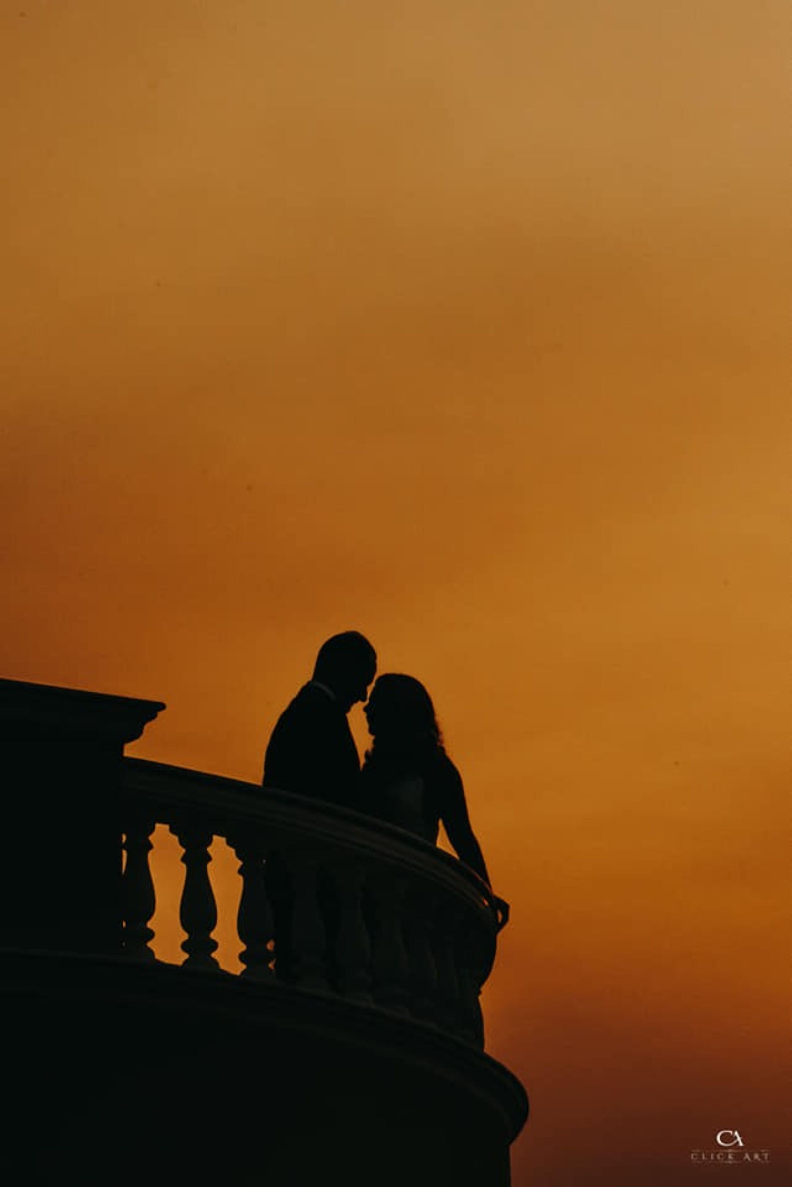 A silhouette of a couple on a balcony against a sunset sky.