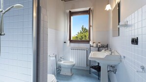 Baño con inodoro, bidé y lavamanos, con azulejos blancos y ventana con vistas a zonas verdes.