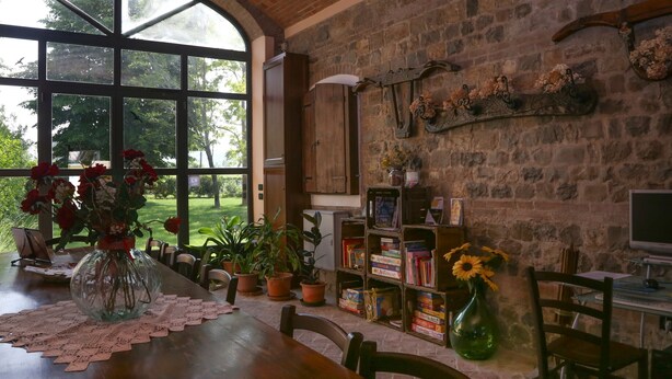 Una habitación acogedora con un amplio ventanal, una mesa de madera con mantel de encaje, plantas en macetas, una estantería y un ordenador sobre un escritorio.
