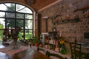 Une pièce chaleureuse avec une grande fenêtre, une table en bois recouverte d’une nappe en dentelle, des plantes en pot, une bibliothèque et un ordinateur posé sur un bureau.