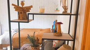 Una habitación con una cama, una estantería metálica negra con varios objetos, una lámpara y una mesita con una vela y una cesta.