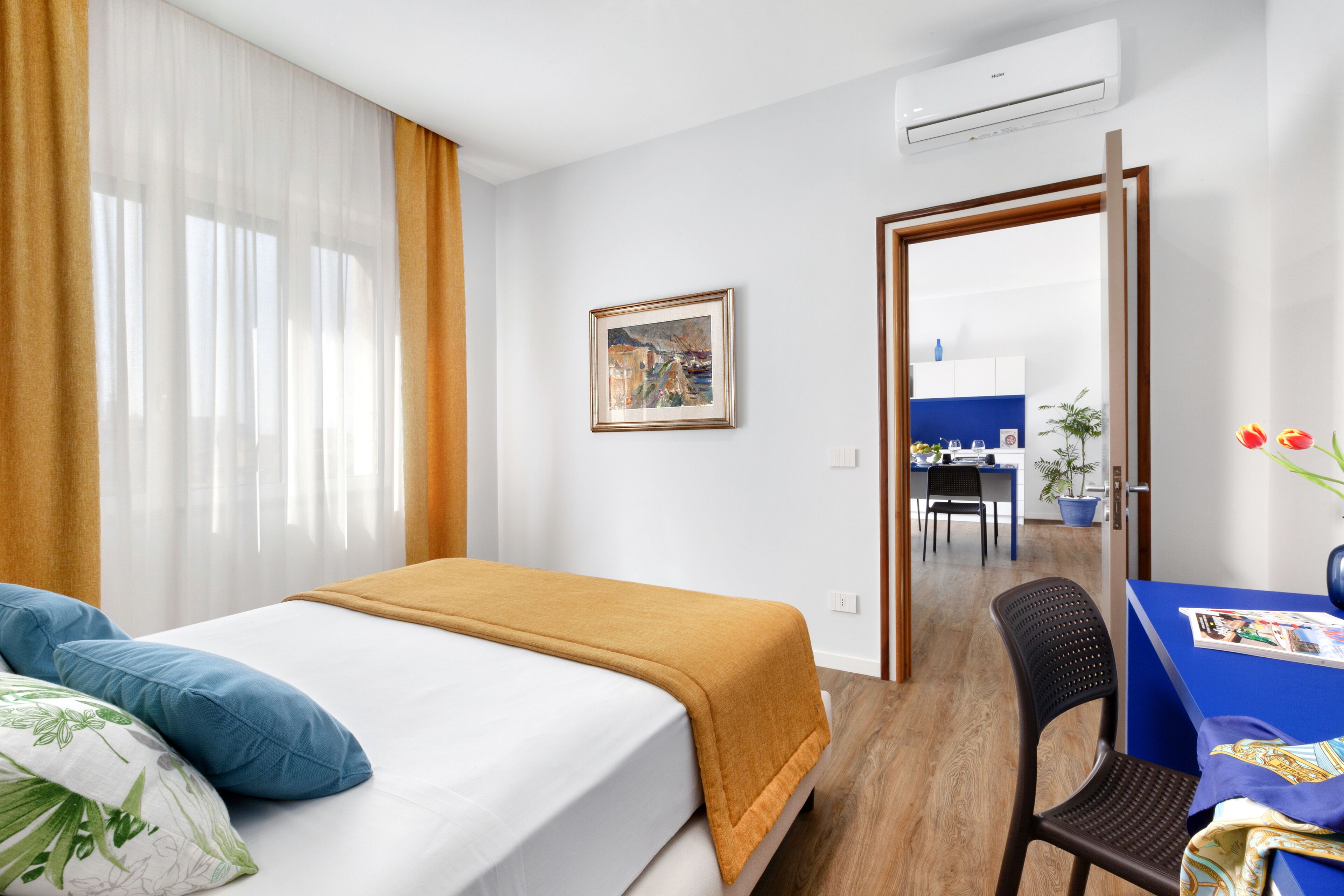 Une chambre d’hôtel avec un lit, un bureau et une chaise. Il y a une fenêtre avec des rideaux, un tableau au mur et un appareil de climatisation.