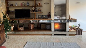Una sala de estar con chimenea, estantes de madera con objetos decorativos, un televisor y un árbol de Navidad.