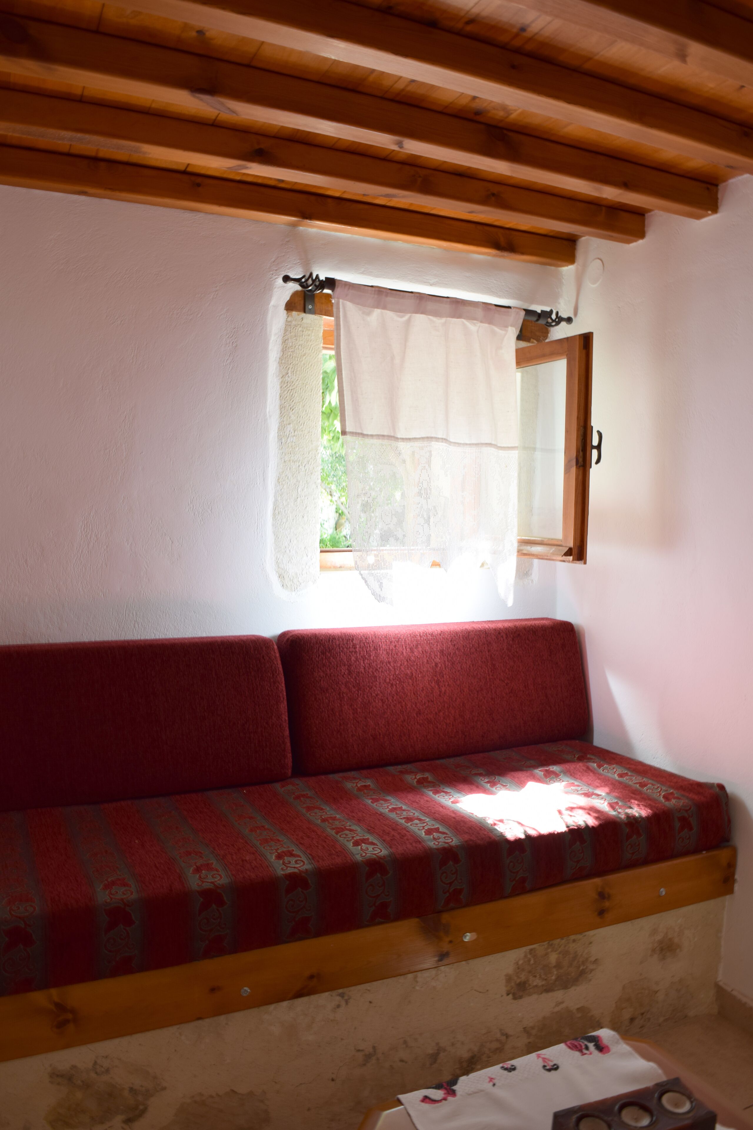 Una habitación con techo de madera, una ventana con cortina, un banco rojo con cojín estampado y una pequeña mesa con mantel.