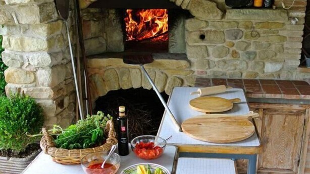 Ein aus Stein gemauerter Pizzaofen im Freien, in dem Feuer brennt, umgeben von einem rustikalen Tisch mit Holzschneidebrettern, einer Schale mit Salat, einer Olivenölflasche und einer Tomatenschale.