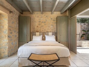 A bedroom with a stone wall, wooden beams, a bed with white linens, and a small folding table. - Villa Nautilus - Family villa with amazing sunset & sea view (Kissamos)