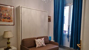 Une pièce avec un canapé marron, une table de chevet blanche, une lampe, un tableau encadré et une fenêtre avec des rideaux bleus.