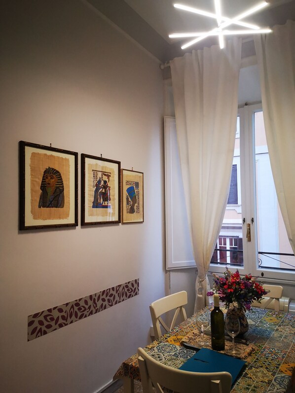 A dining area with a table set for two, framed artwork on the wall, and a window with curtains.