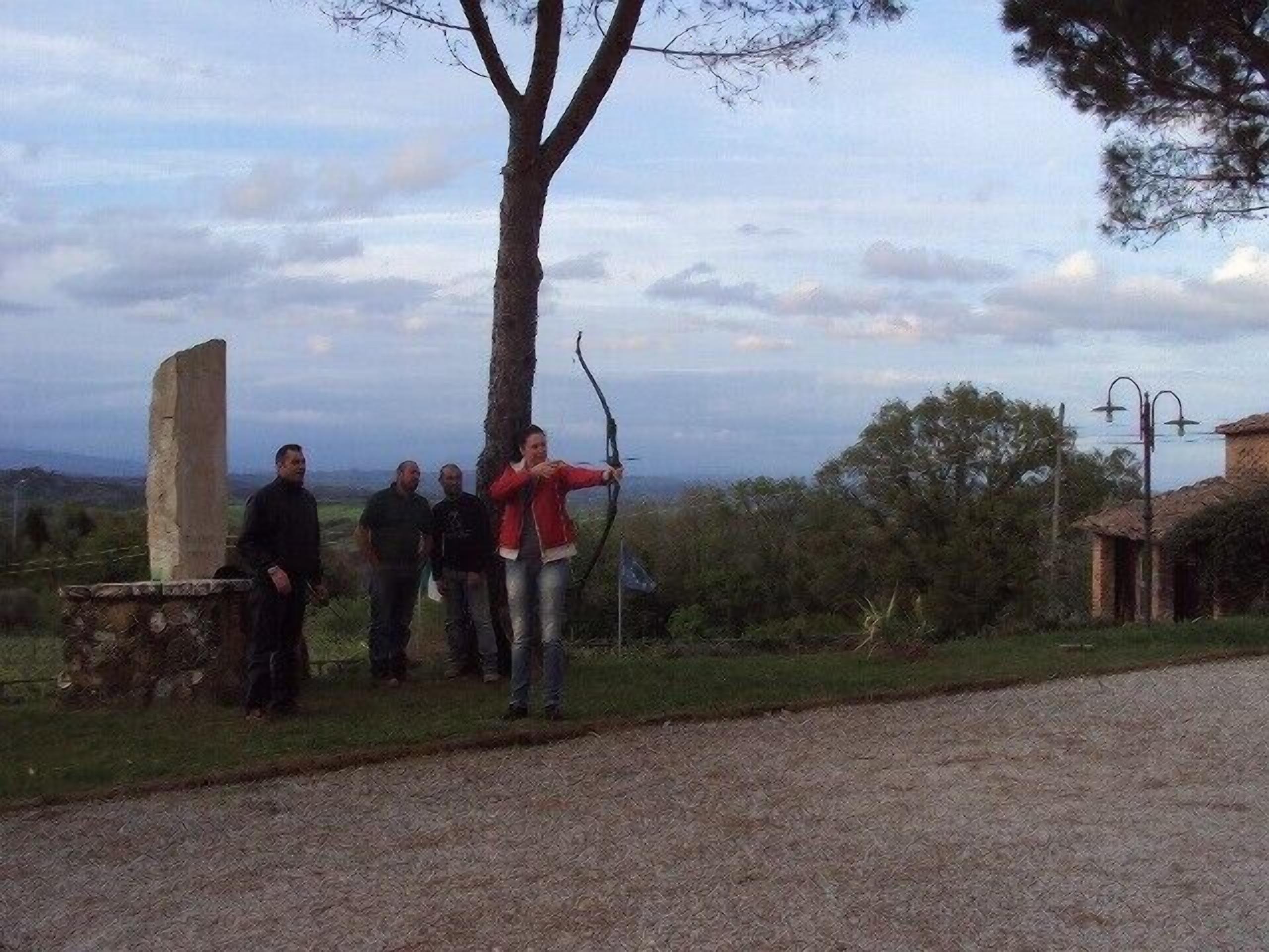 Un gruppo di persone in piedi all'aperto vicino a un albero, con una struttura in pietra e un edificio sullo sfondo.