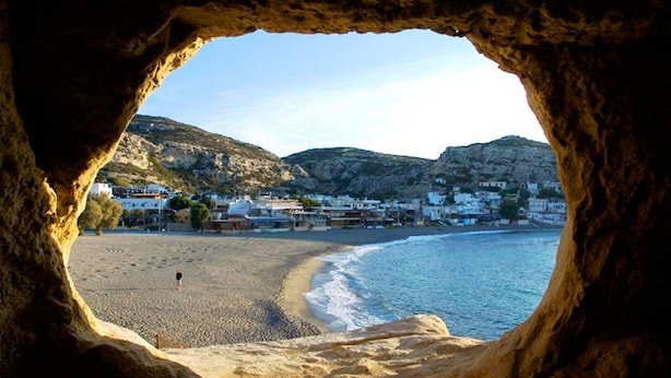 Vista de una playa a través de una abertura en una cueva, con arena, mar tranquilo y edificios al fondo.