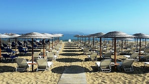 Uma praia com fileiras de espreguiçadeiras e guarda-sóis de palha, que se estendem até o oceano.