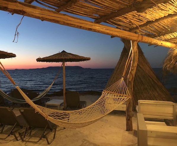 A beach scene with a hammock, chairs, and a thatched roof structure.