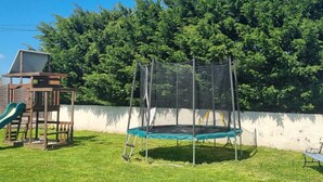 Un parque infantil con un tobogán y un trampolín, rodeado de un césped y un muro bordeado de árboles.