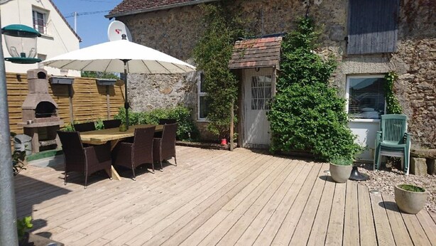 Una terraza de madera con una mesa y sillas de comedor, un edificio de piedra con una antena parabólica y un gran árbol de hoja perenne.