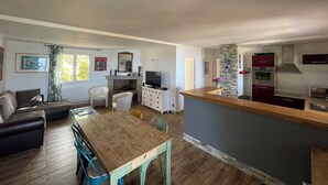 Een ruime woonkamer met een houten eettafel, een bank en een televisie. Een keuken met een steen accentmuur en moderne apparatuur is zichtbaar.