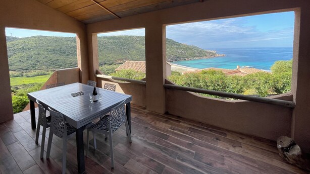 Een kamer met houten plafond, een tafel met stoelen en grote ramen met uitzicht op een strand en heuvels.