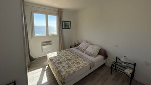 Een slaapkamer met een bed, een nachtkastje, een lamp, een foto aan de muur en een raam met uitzicht op zee.