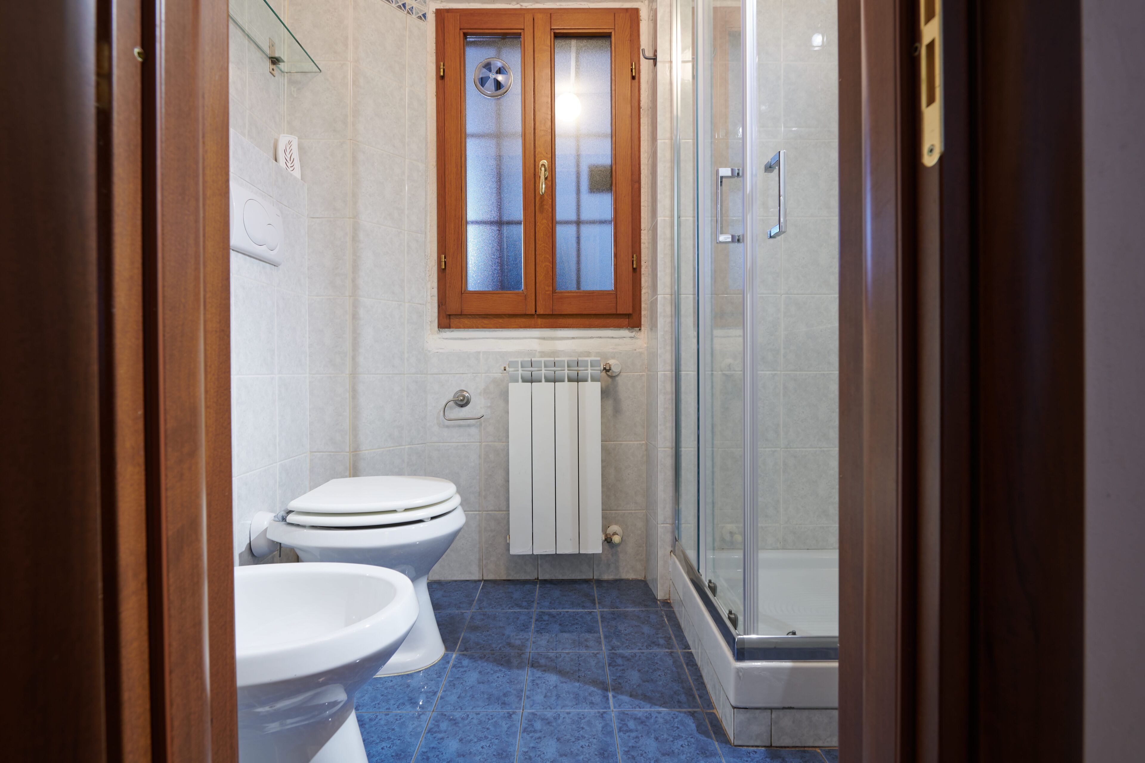 A bathroom with two toilets, a glass shower enclosure, and a wooden-framed window.