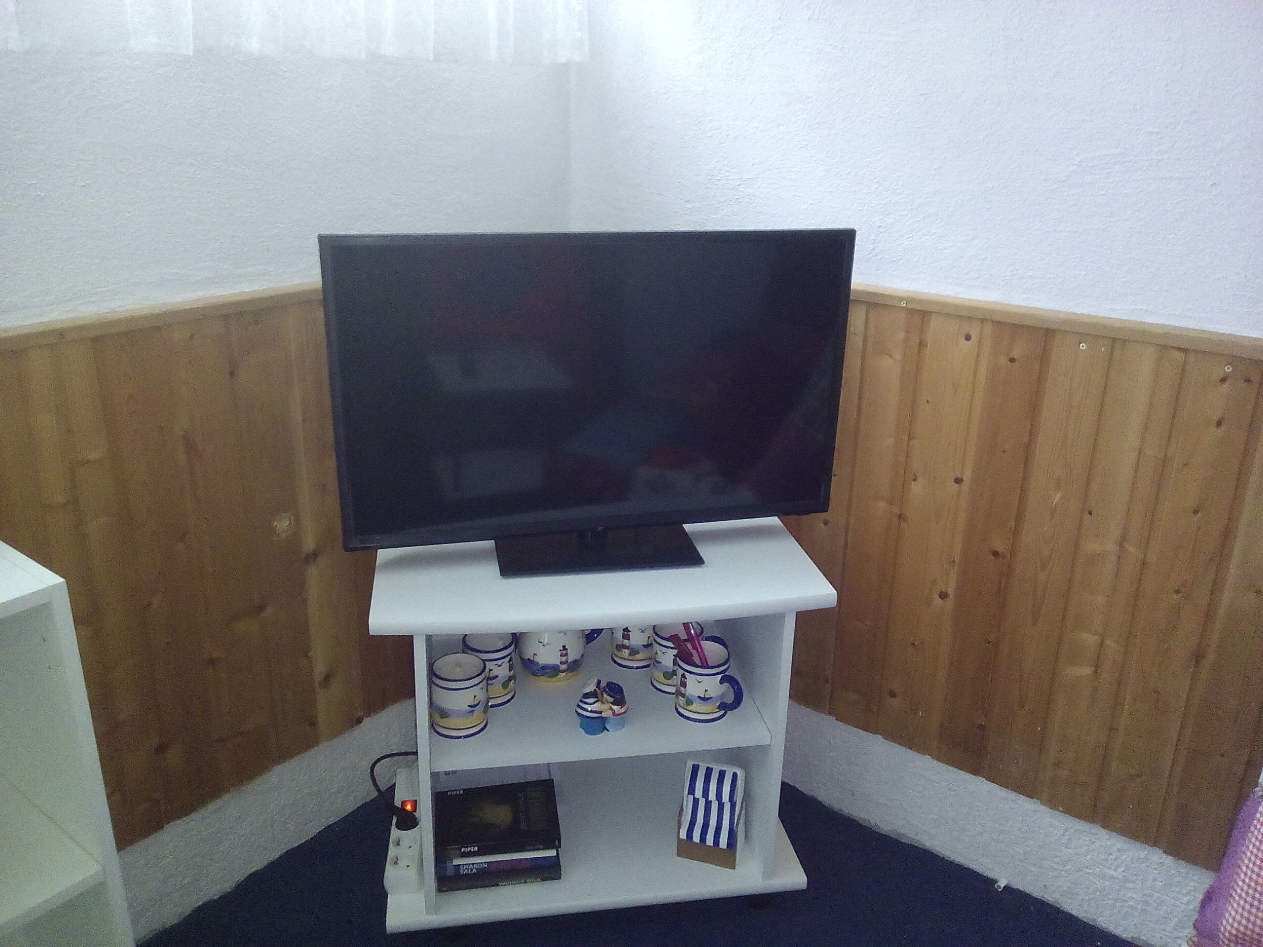 Un téléviseur posé sur un meuble blanc, avec une étagère en dessous où sont rangés divers objets.