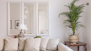 Ein Wohnzimmer mit einem beigefarbenen Ecksofa, einem gemusterten Teppich, einer Topfpflanze und einem Spiegel mit weißem Rahmen.