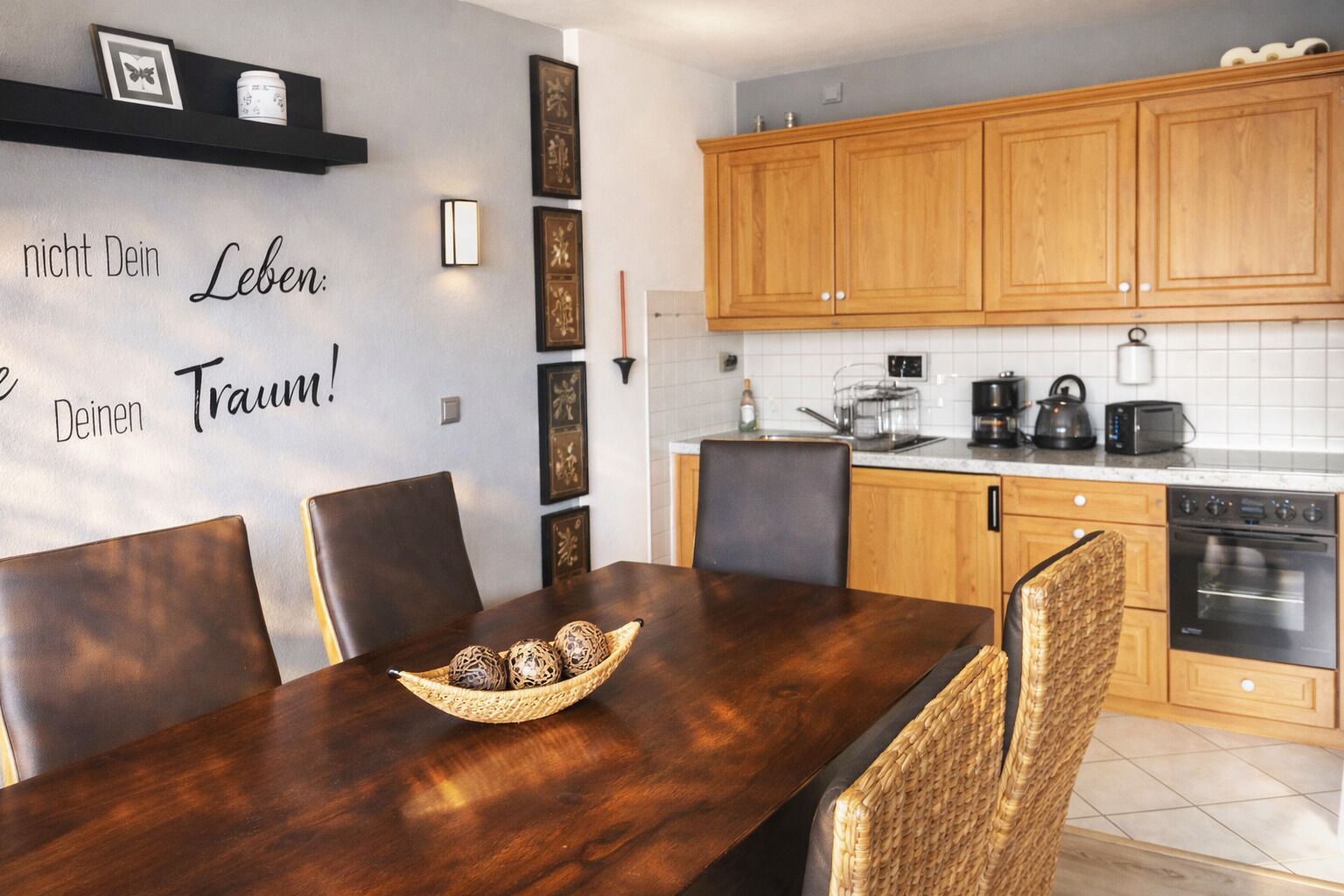 A modern kitchen with wooden cabinets, a dining table with chairs, and a decorative wall with text and framed pictures.
