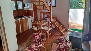 Een compacte keuken met een magnetron, een kleine koelkast en een houten trap naar een balkon. Er staan twee rieten stoelen met bloemenkussens bij een eettafel met stoelen.