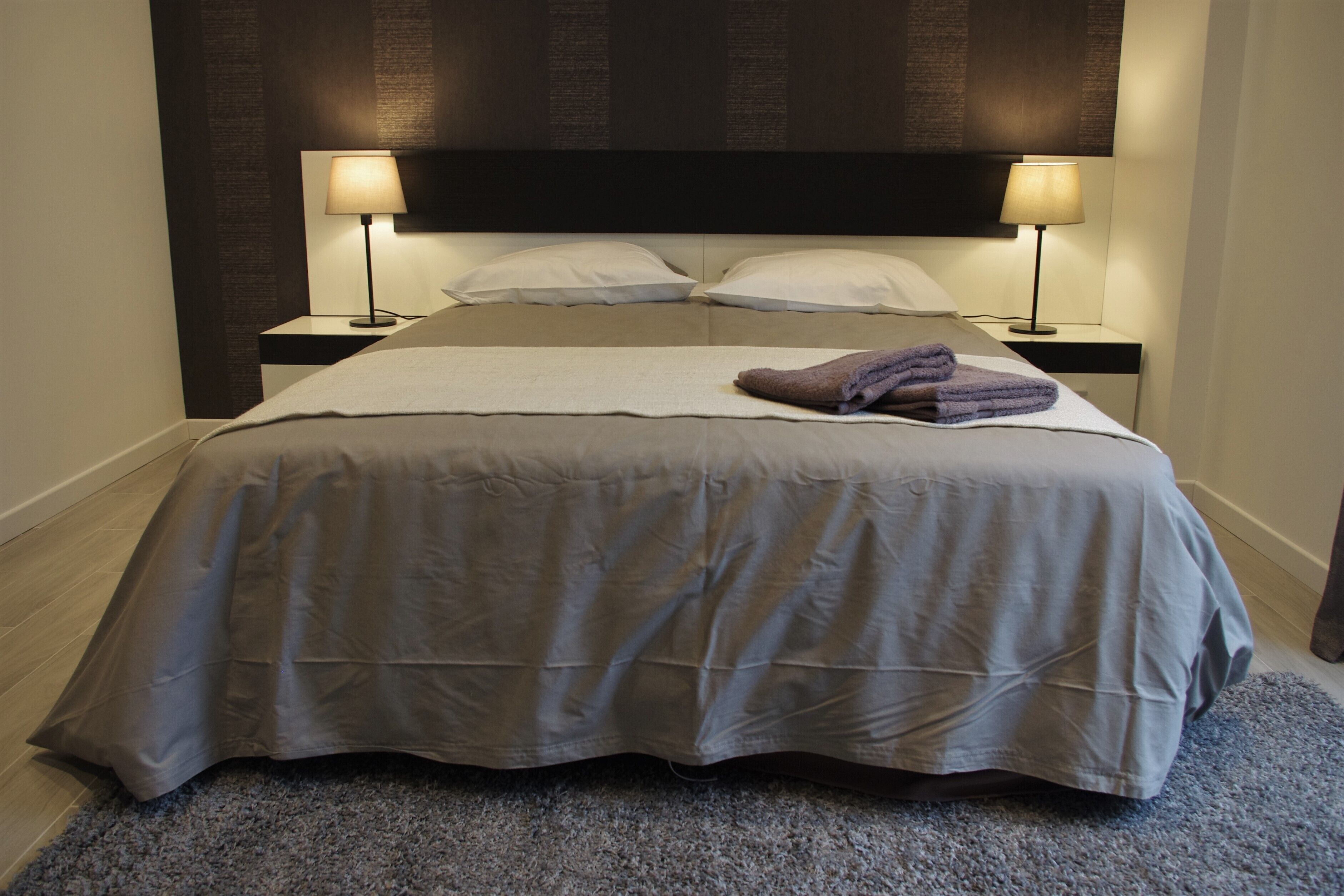 Una cama bien hecha con un edredón gris, dos almohadas blancas y una toalla morada encima. Hay dos lámparas de noche a cada lado de la cama.