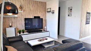 Een moderne woonkamer met een flatscreen televisie, een zwarte leren bank, een witte console en een klein bijzettafeltje met een plant.
