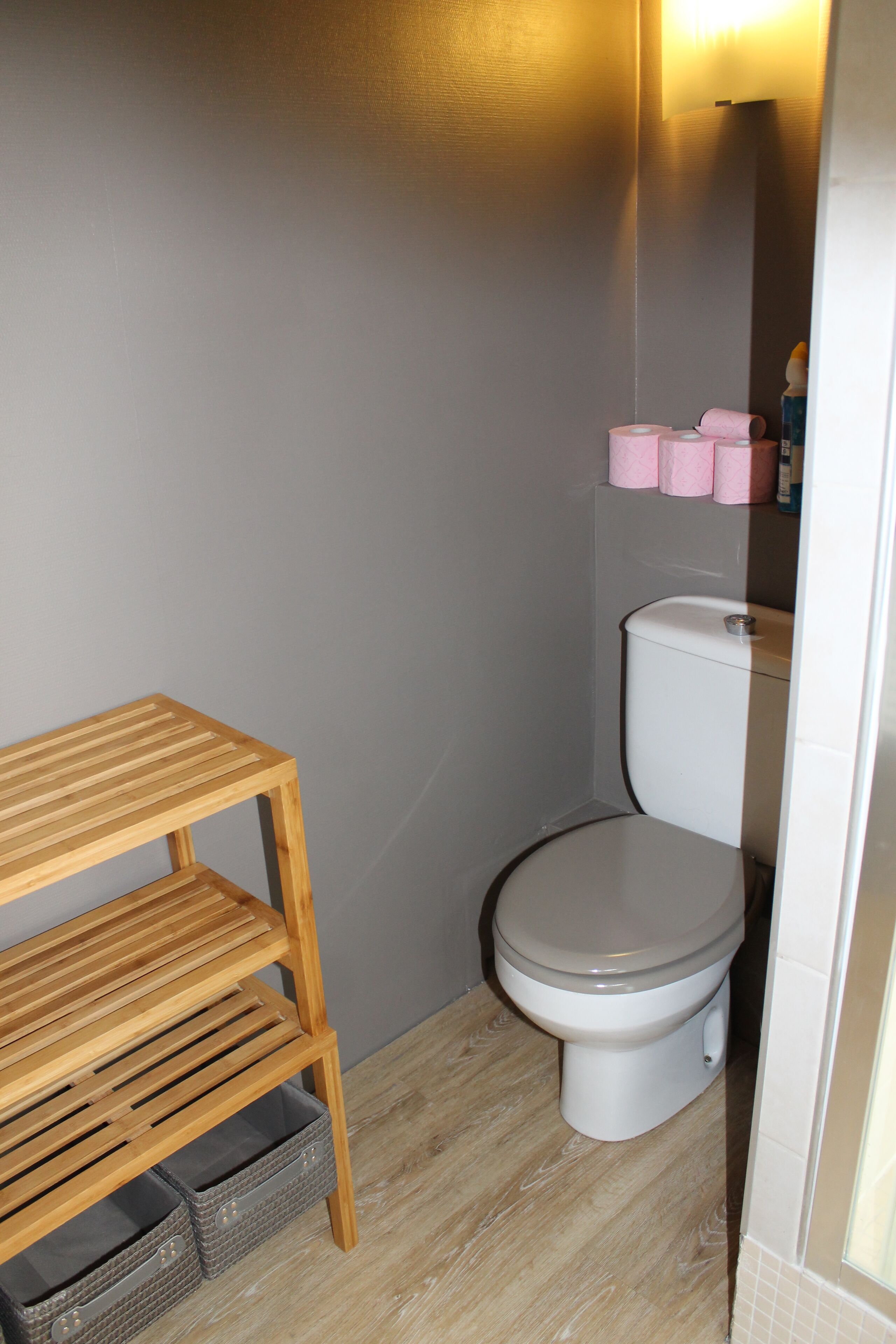 Une petite salle de bain comprenant des toilettes, une étagère en bois et un bac de rangement.