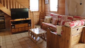 Interior de una cabaña de madera con una televisión sobre un mueble de madera, un sofá con cojines estampados, una mesa de madera y una escalera.