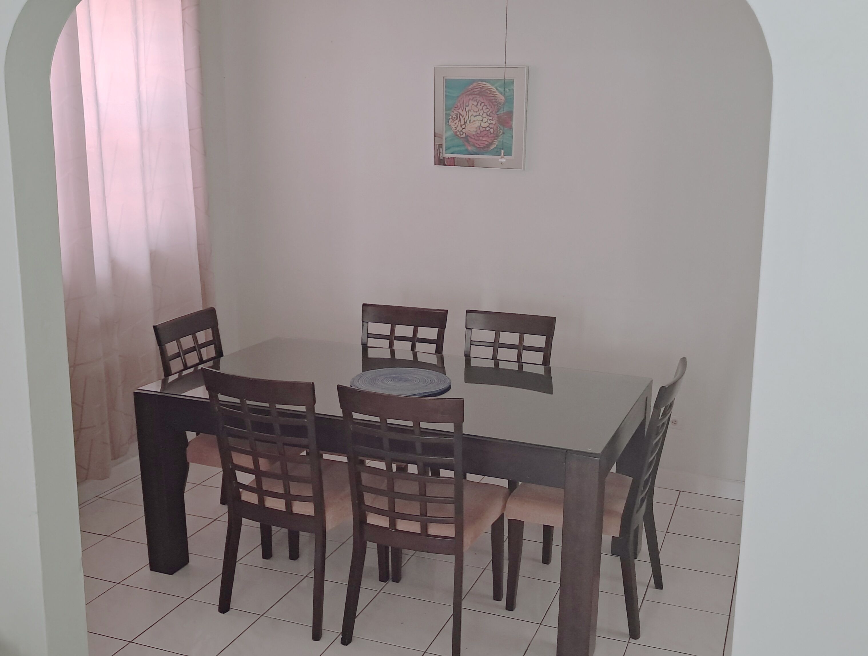 A dining area with a round table and six chairs, a framed picture on the wall, and a window with curtains.