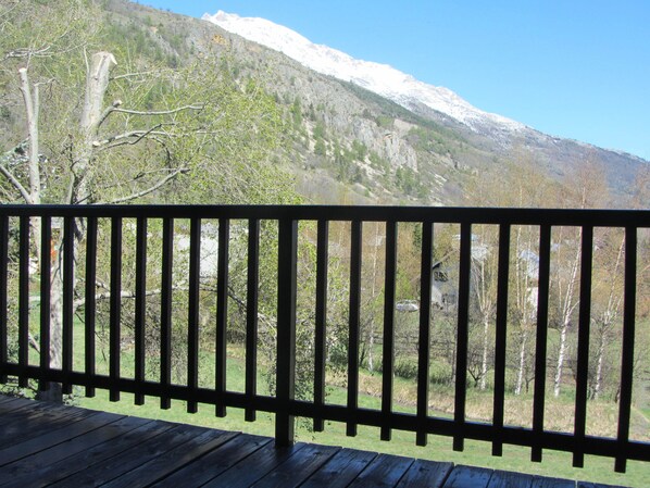 Una terraza de madera con barandilla metálica que da a un paisaje montañoso con árboles y un pico nevado.
