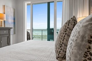 A bedroom with a bed, a nightstand, a lamp, and a view of the ocean through a sliding door.