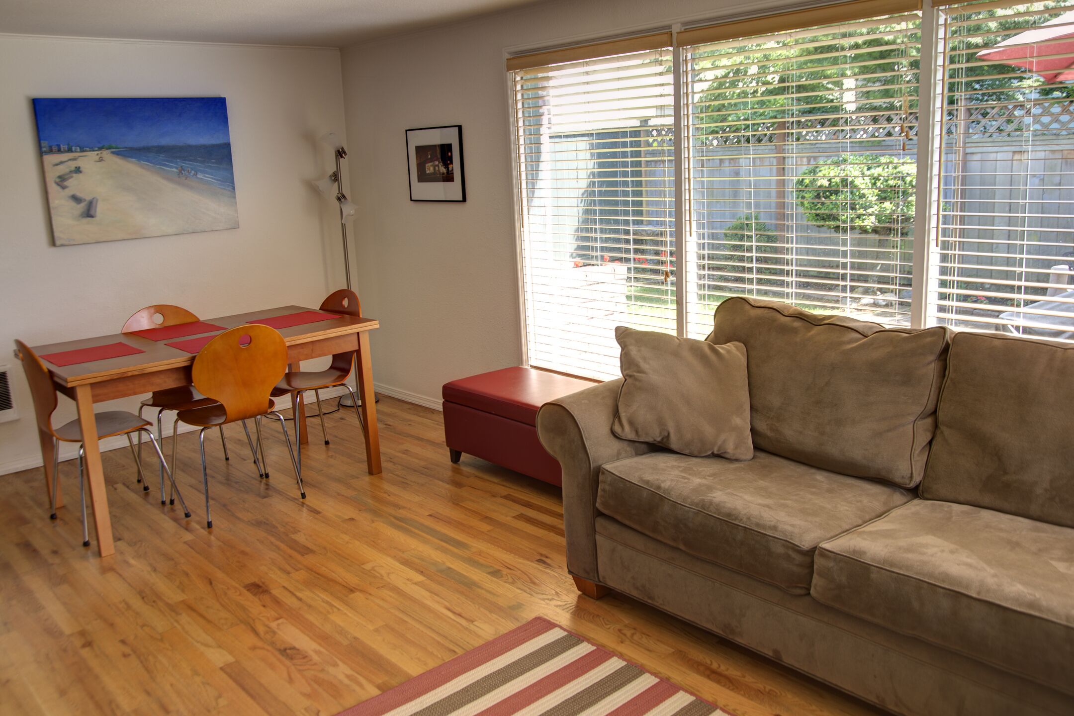 A living room with a wooden floor, a red ottoman, a beige sofa, a dining table with chairs, and a beach painting on the wall.