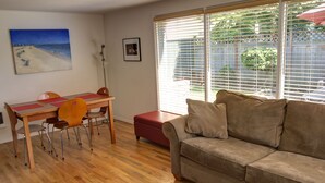 A living room with a wooden floor, a red ottoman, a beige sofa, a dining table with chairs, and a beach painting on the wall.