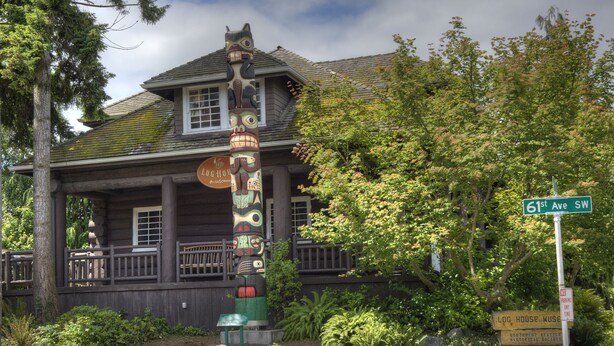 A log house museum with a totem pole and a sign indicating 61st Ave SW.